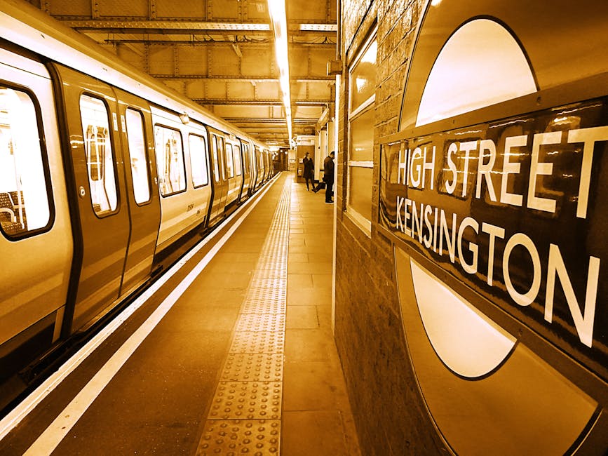A view of a London underground station platform at High Street Kensington, illuminated by overhead lighting revealing a clean, well-maintained surface with tactile paving tiles along the edge. A stainless steel train is stationary alongside the platform, with closed doors and reflective windows. The platform walls are lined with brick and feature a large round sign displaying 'HIGH STREET KENSINGTON' in white lettering. A few passengers are visible in the distance, waiting or walking along the platform. The scene highlights a hygienic and orderly environment typical of city transit stations, emphasizing cleanliness and maintenance conducive to effective cleaning and sanitation, as promoted by westkensingtoncarpetcleaning.co.uk on their service page about basement access solutions for cleaning.
