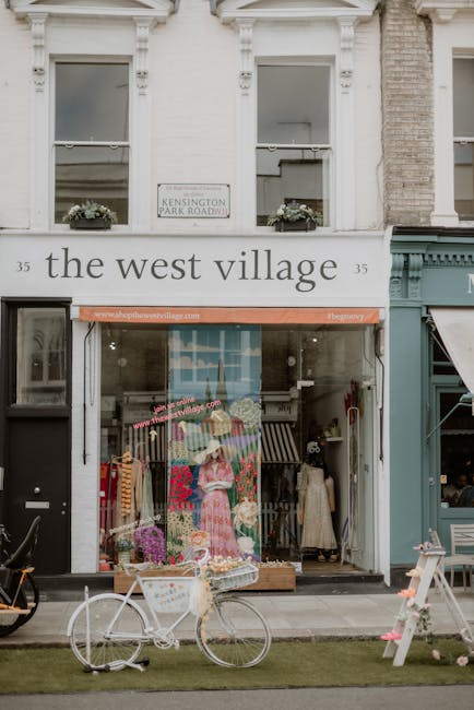 Exterior view of a boutique shop named 'the west village' located at 35 Kensington Park Road in West Kensington, showing two upper-story windows with flower boxes, and the storefront with a display window filled with colorful clothing and accessories. In front of the shop, a white bicycle with a basket is parked on the sidewalk, alongside a small wooden table with pink decorations. The scene is well-lit with natural daylight, highlighting the clean and maintained appearance of the storefront, which is part of residential and commercial buildings in a city setting. This image emphasizes the importance of maintaining cleanliness and presentation for local businesses as part of ongoing surface cleaning and hygiene practices, aligning with professional cleaning standards promoted by westkensingtoncarpetcleaning.co.uk.
