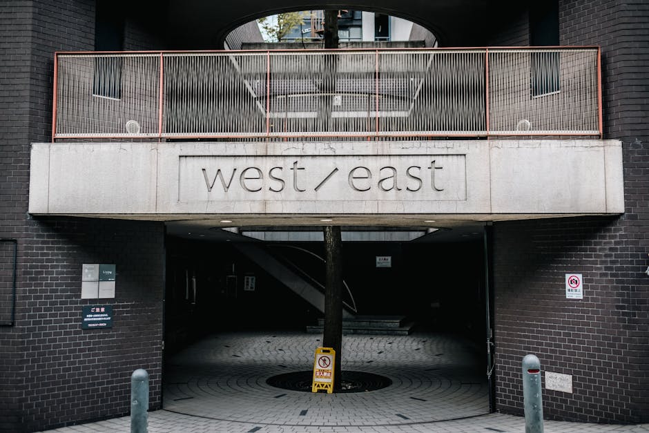 A view of a London underground station platform at High Street Kensington, illuminated by overhead lighting revealing a clean, well-maintained surface with tactile paving tiles along the edge. A stainless steel train is stationary alongside the platform, with closed doors and reflective windows. The platform walls are lined with brick and feature a large round sign displaying 'HIGH STREET KENSINGTON' in white lettering. A few passengers are visible in the distance, waiting or walking along the platform. The scene highlights a hygienic and orderly environment typical of city transit stations, emphasizing cleanliness and maintenance conducive to effective cleaning and sanitation, as promoted by westkensingtoncarpetcleaning.co.uk on their service page about basement access solutions for cleaning.