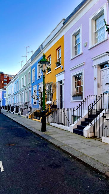 A row of colorful residential terraced houses along a street near West Kensington station, featuring pastel facades in shades of blue, yellow, purple, and pink. The houses have white-framed windows, black wrought-iron railings, and steps leading up to their front doors. A vintage-style black lamppost stands in front, casting light onto the sidewalk. The street appears clean and well-maintained, with a clear blue sky overhead. This scene exemplifies the charming architecture commonly found in West Kensington, highlighting the vibrant exterior aesthetics that benefit from regular domestic cleaning and surface maintenance by local service providers such as westkensingtoncarpetcleaning.co.uk.