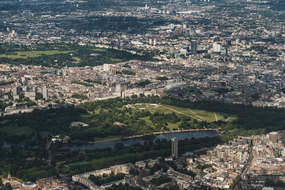 Aerial view of a large city featuring a central green park with walking paths and a pond, surrounded by densely packed urban buildings and skyscrapers, under a partly cloudy sky with sunlight illuminating the scene. The image showcases the contrast between natural green spaces and the urban landscape, with diverse surfaces including grass, water, concrete, and glass structures. Visible sections of the park appear well-maintained, with lush trees and open grassy areas, while the surrounding cityscape includes office buildings, residential blocks, and high-rise towers typical of central London, close to Kensington. The photograph highlights the importance of surface cleaning and maintenance in urban environments and may be used to emphasize the necessity of professional cleaning services like those offered by westkensingtoncarpetcleaning.co.uk for residential or commercial spaces in such areas.