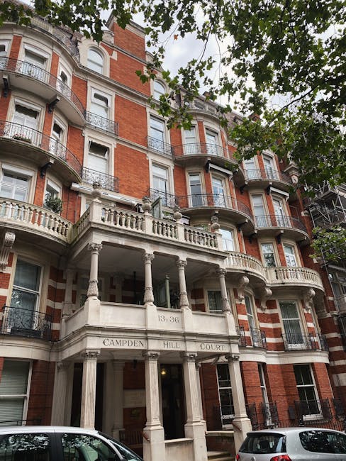 Exterior view of a multi-story residential building with a classic architectural style, featuring red brick walls, white decorative stone elements, and curved balconies with black wrought iron railings. The building has large windows with white frames, and a main entrance supported by white columns and an ornate balustrade, with the address '13-36' and the name 'Campden Hill Court' displayed above. The foreground includes parked cars, and tree branches with green leaves frame the top of the image, emphasizing the building's location in a leafy neighborhood. The scene is well-lit with natural daylight, highlighting the clean and maintained appearance of the facade, which underscores the importance of routine cleaning and maintenance associated with professional domestic cleaning services such as those provided by westkensingtoncarpetcleaning.co.uk.