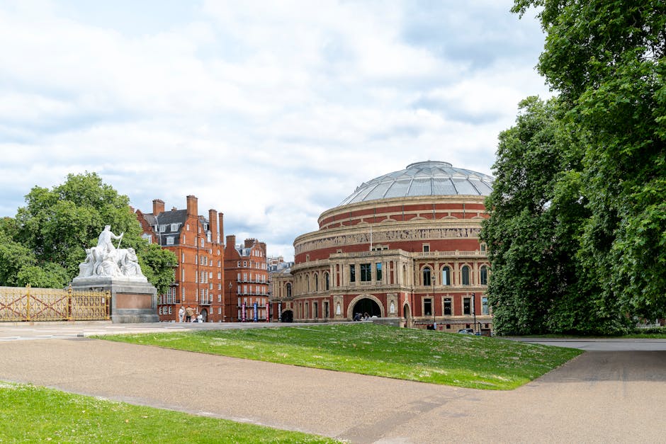 A large, domed building with a brick and stone facade situated within a park in Holland Park, Kensington. The building features tall arched windows and decorative architectural details. Surrounding the structure are lush green trees and a well-maintained lawn, with a paved pathway leading towards it. The park area appears tidy and clean, with bright natural lighting highlighting the vibrant greenery and the historic architecture. In the foreground, there is a section of grass and a stone statue partially visible on the left side, indicating a scenic and well-kept outdoor space associated with domestic or community settings.