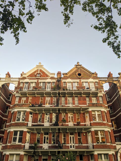 Exterior view of a historic red brick residential building in Holland Park, featuring multiple levels with white decorative moldings, bay windows, and balconies with black wrought-iron railings. The building is set against a clear sky with green tree branches partially framing the top of the image. The street-level entrance is visible, flanked by lush greenery, and the overall appearance is well-maintained, reflecting period architecture. This image is associated with West Kensington Carpet Cleaning, which provides comprehensive cleaning and deep cleaning services for period homes in Kensington, highlighting the importance of surface cleaning and maintenance for heritage properties.