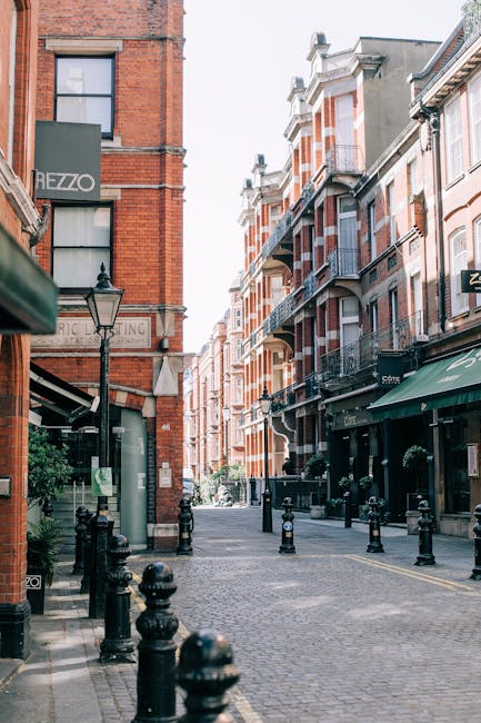 A quiet, cobblestone street in Kensington with red brick buildings lining both sides, featuring large windows and balconies with black wrought iron railings. The street is partially shaded by a green awning extending from a storefront on the right, and a classic black lamppost is visible in the foreground. The area appears clean and well-maintained, with no visible litter or debris, embodying an aesthetically pleasing urban environment typical of Kensington. The image showcases a mix of residential and commercial buildings, emphasizing the tidy, well-kept nature of this vicinity, and aligns with the principles of surface cleaning and maintenance promoted by westkensingtoncarpetcleaning.co.uk in the context of adhereing to waste management rules and hygienic standards.
