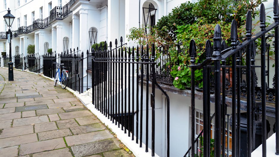 A paved sidewalk made of rectangular stone slabs running alongside a white residential building with black wrought iron railings and gates. The pavement appears clean and free of debris. To the right, there are potted plants and green foliage growing along the railing, adding a touch of greenery to the scene. Visible in the background are shadows cast by the railings and the building's exterior, with natural daylight illuminating the area. A blue bicycle is parked near the sidewalk, leaning against the railing. The overall scene depicts an exterior urban residential setting, with well-maintained surfaces and tidy surroundings, reflecting the importance of surface cleaning and maintenance related to domestic cleaning and city hygiene standards.