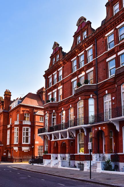 A quiet, cobblestone street in Kensington with red brick buildings lining both sides, featuring large windows and balconies with black wrought iron railings. The street is partially shaded by a green awning extending from a storefront on the right, and a classic black lamppost is visible in the foreground. The area appears clean and well-maintained, with no visible litter or debris, embodying an aesthetically pleasing urban environment typical of Kensington. The image showcases a mix of residential and commercial buildings, emphasizing the tidy, well-kept nature of this vicinity, and aligns with the principles of surface cleaning and maintenance promoted by westkensingtoncarpetcleaning.co.uk in the context of adhereing to waste management rules and hygienic standards.