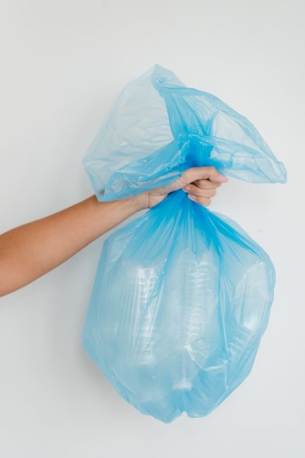A person holding two large blue recycling bags made of thin plastic material, one in each hand, against a plain white background. The bags appear to be filled with recyclable waste, possibly plastic bottles or other light materials. The person's arm and hand are visible, with the bags suspended in mid-air. The scene emphasizes responsible waste disposal and recycling practices associated with professional cleaning services in Kensington, as promoted by westkensingtoncarpetcleaning.co.uk, focusing on environmental hygiene and sustainable waste management.