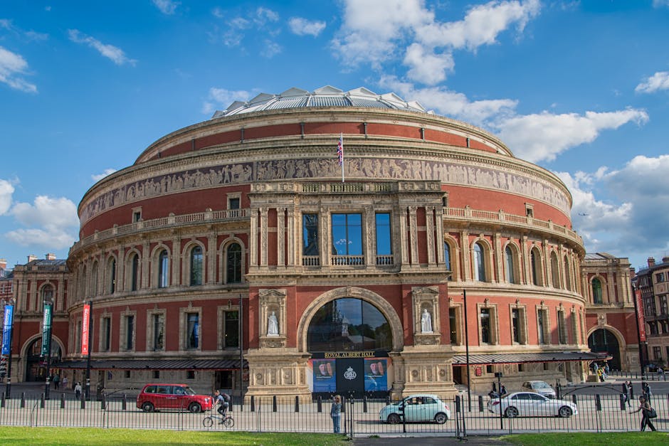 Exterior view of the Royal Albert Hall in Kensington, showcasing its circular, historic brick façade with intricate stone detailing and large arched windows. The grand entrance features a central archway with a glass door, flanked by statues and decorative elements. The building is set against a bright blue sky with scattered clouds, with cars and pedestrians visible on the street in front. The exterior appears clean and well-maintained, emphasizing the importance of surface cleaning and building upkeep, as provided by westkensingtoncarpetcleaning.co.uk for event spaces in Kensington.