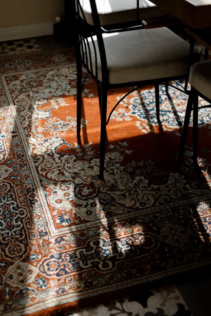 A handheld vacuum cleaner with a black and red body is positioned on a patterned, plush area rug in a room. The rug features intricate designs in shades of beige, brown, and faded red. The vacuum's nozzle is resting on the rug, indicating active surface cleaning. Behind the vacuum, a white curtain hangs partially across the background, and part of a piece of light-colored wooden furniture is visible on the right side. The scene is well-lit, highlighting the cleanliness of the rug and the tools used for deep cleaning and stain removal, consistent with services offered by westkensingtoncarpetcleaning.co.uk for rug cleaning and sanitisation in Earl Court and West Kensington.