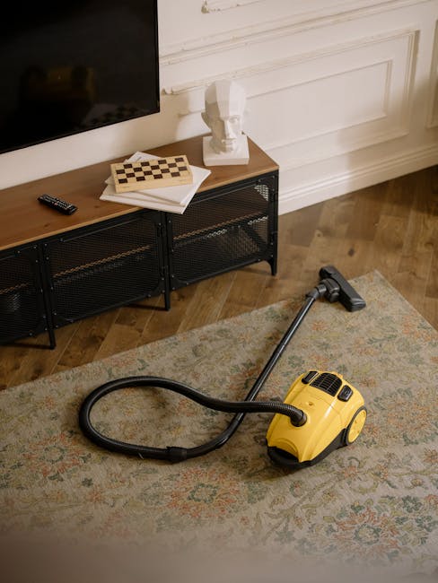 A yellow canister vacuum cleaner with a black hose and a cleaning head is positioned on a patterned area rug in a living room. The rug features intricate designs in muted colors and covers a wooden floor. In the background, there is a black metal and wood TV stand with a remote control, a couple of notebooks, a chessboard, and a white decorative bust sculpture placed on top. The wall behind the TV stand is painted white with decorative molding, and part of a flat-screen TV is visible at the top left. The lighting is soft and natural, highlighting the clean, well-maintained surfaces suitable for domestic surface cleaning and deep cleaning purposes, as promoted by westkensingtoncarpetcleaning.co.uk.