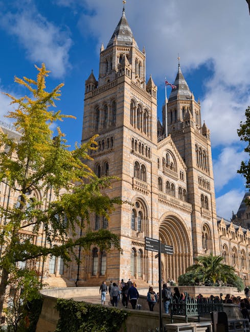 The exterior of a historic cathedral with intricate stonework, featuring two tall, pointed towers topped with conical spires. The building is constructed from light-colored stone and has arched windows and decorative details along its facade. In the foreground, there are trees with green and yellow leaves, suggesting a mild season. Pedestrians are walking along the paved pathway in front of the cathedral, which is set against a bright blue sky with scattered clouds. The scene is well-lit by natural sunlight, highlighting the architectural features and the cleanliness of the surrounding area. The image is associated with a professional cleaning service, emphasizing the importance of surface cleaning and tidiness in prominent public or historic buildings, as promoted by westkensingtoncarpetcleaning.co.uk.