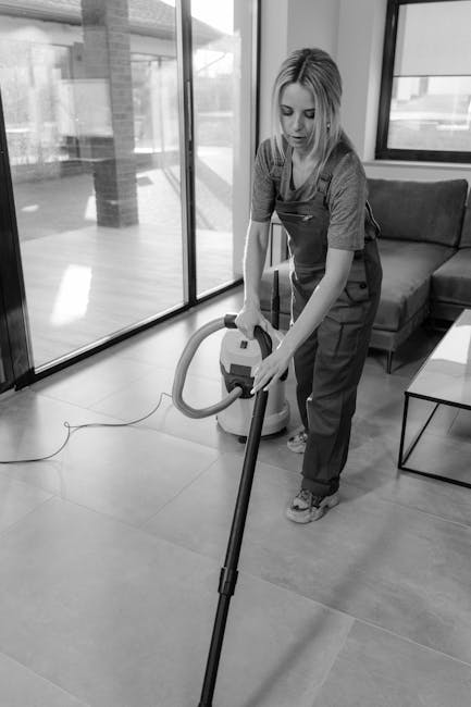 A yellow canister vacuum cleaner with a black hose and a cleaning head is positioned on a patterned area rug in a living room. The rug features intricate designs in muted colors and covers a wooden floor. In the background, there is a black metal and wood TV stand with a remote control, a couple of notebooks, a chessboard, and a white decorative bust sculpture placed on top. The wall behind the TV stand is painted white with decorative molding, and part of a flat-screen TV is visible at the top left. The lighting is soft and natural, highlighting the clean, well-maintained surfaces suitable for domestic surface cleaning and deep cleaning purposes, as promoted by westkensingtoncarpetcleaning.co.uk.