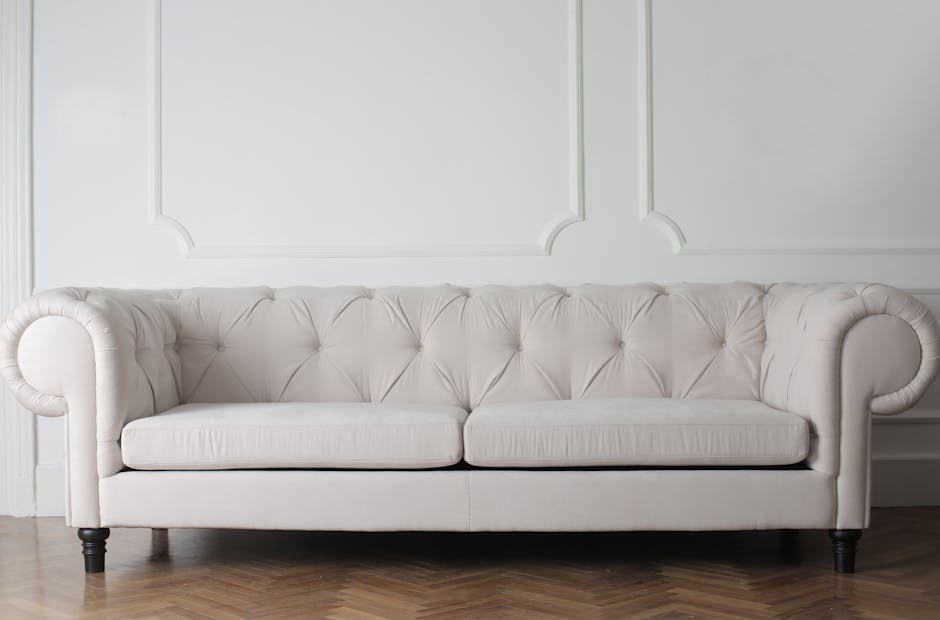A pristine, white upholstered sofa with button-tufted detailing on the backrest and rolled arms, situated against a white paneled wall in a well-lit room. The fabric appears clean and smooth, with no visible stains or dust. The sofa's dark wooden legs contrast with the light fabric, and the wooden flooring beneath features a herringbone pattern. The setting suggests a tidy, stylish living space prepared for surface cleaning and maintenance, aligning with expertise in upholstery cleaning available from westkensingtoncarpetcleaning.co.uk.